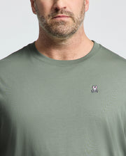 A man models a light green Psycho Bunny BIG AND TALL CLASSIC CREW NECK TEE, made from 100% Pima cotton. It features a small white rabbit logo above crossed bones embroidered on the chest. The focus is on the tee's upper portion against a plain background.