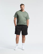 A man, with a neutral expression, stands against a plain background wearing Psycho Bunny's BIG AND TALL CLASSIC CREW NECK TEE in green, paired with black shorts. Made from 100% Pima cotton, the tee ensures comfort. He complements his look with white socks and sneakers while resting his hands in his pockets.