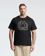 A bearded man with short hair wears an eco-friendly black Psycho Bunny t-shirt made from Peruvian Pima cotton. The white graphic features a bunny head with crossbones, and the text "Psycho Bunny" is displayed. He's paired with beige pants.