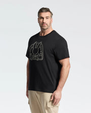 A bearded man with short hair wears Psycho Bunny's MENS BIG AND TALL MELBOURNE GRAPHIC TEE, a black eco-friendly Peruvian Pima cotton shirt featuring a bunny and crossbones design. He pairs it with beige pants, standing against a plain background and looking at the camera.