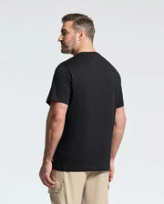 A man with short hair and a beard stands slightly away, wearing a Psycho Bunny MENS BIG AND TALL MELBOURNE GRAPHIC TEE in black, made of eco-friendly Peruvian Pima cotton, and beige cargo pants. The background is light gray.