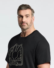 A man with short hair and a beard wears the Psycho Bunny Men's Big and Tall Melbourne Graphic Tee, crafted from eco-friendly Peruvian Pima cotton. The black T-shirt showcases a bunny with crossbones and "PSYCHO BUNNY" text. The background is plain and light-colored.