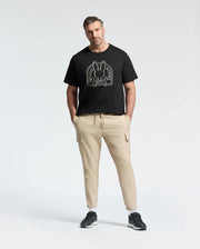 A man stands against a white background wearing Psycho Bunny's eco-friendly black "Melbourne Graphic Tee" in soft Peruvian Pima cotton featuring a bunny graphic, paired with beige cargo pants and black sneakers, hands in pockets.