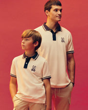 An adult and child wear matching Psycho Bunny MENS WADE PIQUE POLO shirts (MT0100044) in Pima cotton, featuring the embroidered bunny logo, standing against a plain coral background and looking off to the side with relaxed expressions.