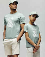 Two people wear matching light green Psycho Bunny MENS DEREK CENTER GRAPHIC TEEs embroidered with a white Bunny and crossbones. The adult pairs it with light shorts, hands in pockets; the child stands arms crossed against a neutral background.