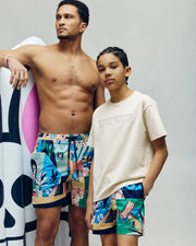 Two people pose together; one is shirtless in colorful Psycho Bunny MENS BLAISE SWIM TRUNK - MB1000028, and the other wears a beige Psycho Bunny tee and shorts. Bunny logos are visible, with a large inflatable bunny skull in the background.