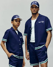 A boy and a man wear matching Psycho Bunny MENS BASIL TAPED CAMP COLLAR SHIRTS (MT0500004) in navy blue cotton-modal, paired with white and green-trimmed shorts and caps, confidently posing against a plain background.