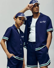 Two people wear matching Psycho Bunny navy KIDS BASIL TAPED CAMP COLLAR shirts and shorts, made from a breathable, flexible premium cotton-modal blend. The adult rests his arm on the boy’s shoulder.