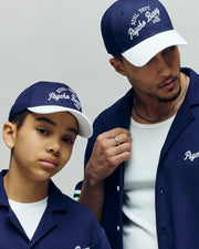 An adult and child wear matching navy blue Psycho Bunny hats and shirts in varsity style. The adult adjusts his KIDS MATTEO BASEBALL CAP (KO1500002) while the child faces the camera, both calm against a plain background.
