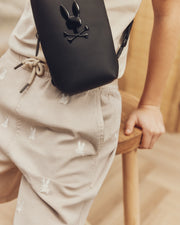 Sporting Psycho Bunny's KIDS ALL OVER WILLIS STRETCH TENCEL SHORT, adorned with white bunny patterns, paired with a crisp white shirt and a crossbody bag flaunting a bunny and crossbones logo. The indoor casual setting enhances their regular fit style effortlessly.