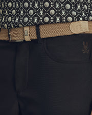 Close-up of a waist in dark pants with a skull emblem, featuring Psycho Bunny's MENS STRETCH BRAIDED BELT - B6A890F200 in beige with a rabbit logo. The ensemble is completed by a patterned shirt with tennis balls and rabbits for an eye-catching look.