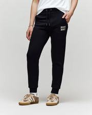 A child models the Psycho Bunny KIDS WADE CORD PATCH SWEATPANT (KB0800002), featuring an embroidered wordmark on the left thigh, paired with a white shirt and beige sneakers against a plain light background for a classic athletic look.