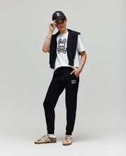 A person poses against a plain background, wearing a black cap, a white Psycho Bunny T-shirt with a bold cartoon bunny graphic, KIDS WADE CORD PATCH SWEATPANT - KB0800002 by Psycho Bunny, and tan Adidas sneakers.