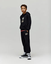 A boy with short curly hair stands against a plain background wearing Psycho Bunny KIDS WADE CORD PATCH SWEATPANT (KB0800002), a black hoodie with a bunny graphic, and black high-top sneakers, hands in his pockets.