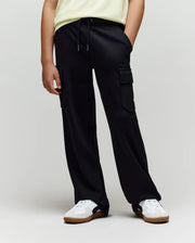 A person wearing a light yellow shirt, Psycho Bunny KIDS NASH CARGO SWEATPANT - KB0800004, and white sneakers with brown soles stands with hands in pockets against a plain background, blending street style and sweatpant comfort.