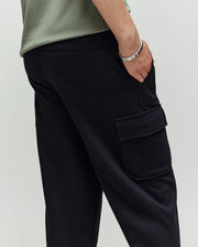A person wears Psycho Bunny KIDS NASH CARGO SWEATPANT - KB0800004 in black with cargo pockets and a light green shirt, one hand in their pocket and a silver bracelet on their wrist—blending style with sweatpant comfort.