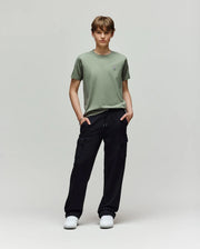 A young person faces forward, wearing a light green t-shirt, white sneakers, and Psycho Bunny KIDS NASH CARGO SWEATPANT - KB0800004 with cargo pockets, hands in pockets, enjoying sweatpant comfort against a plain light gray background.