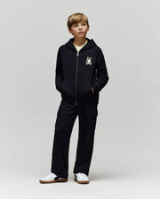 A boy stands with hands in pockets, wearing a black zip-up hoodie with a cartoon chest print, Psycho Bunny KIDS NASH CARGO SWEATPANT - KB0800004 with cargo pockets, and white sneakers, against a plain light grey background.