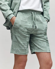 Teen | A person wears Psycho Bunny's KIDS KELVIN TOWEL TERRY SWEATSHORT (KB0900002) and a matching jacket in sage green jacquard terry, both embossed with a checkered bunny pattern featuring repeating Bugs Bunny faces, hand in pocket.