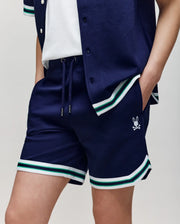 Teen | A child wears Psycho Bunny’s KIDS BASIL TAPED SHORT (KB0900003) in a premium cotton-modal blend, featuring an embroidered Bunny Drawstring and crossbones logo on one leg, paired with a navy shirt with white and green striped trim. One hand rests in their pocket.