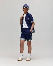 Teen | A person poses against a plain background wearing Psycho Bunny’s KIDS BASIL TAPED SHORT - KB0900003 with white and green trim, a white cotton-modal T-shirt, white sneakers, and a navy cap. Their hand rests on their collar as they face the camera.