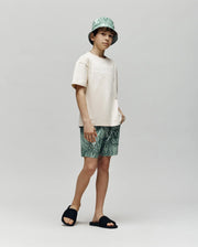 Kids | A young boy stands against a plain background in Psycho Bunny's KIDS CHUCK ALL OVER PRINT LIGHTWEIGHT SWIM TRUNK (KB1000002), paired with a light beige tee, matching green bucket hat, and black slides, gazing to the side with a relaxed look.