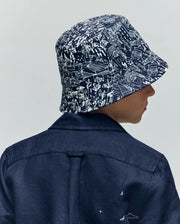 A child is seen from behind wearing a dark blue jacket and the Psycho Bunny KIDS NAXOS REVERSIBLE BUCKET HAT (KO1500001), featuring a white abstract pattern, set against a plain light background.