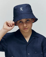 A young boy wearing the Psycho Bunny KIDS NAXOS REVERSIBLE BUCKET HAT (KO1500001), featuring a navy design with an embroidered bunny and crossbones logo, looks at the camera while holding his hat’s brim.