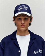 A young person sports a Psycho Bunny KIDS MATTEO BASEBALL CAP (KO1500002) in navy cotton twill with an adjustable strap, teamed with a matching jacket over a white t-shirt. Both the cap and jacket feature bold Psycho Bunny branding.