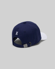 The Psycho Bunny KIDS MATTEO BASEBALL CAP (KO1500002) features a varsity-style navy blue cotton twill with a white brim, small white embroidered logo above the adjustable metal buckle strap, shown from the back on a light gray background.