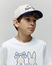 A young boy with curly brown hair wears the Psycho Bunny KIDS MATTEO BASEBALL CAP (KO1500002) in cream and navy cotton twill with an adjustable strap, paired with a white T-shirt featuring a cartoon bunny and crossbones, against a plain background.