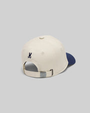 The KIDS MATTEO BASEBALL CAP (KO1500002) by Psycho Bunny is a cream cotton twill cap with a navy brim, seen from the back. It has an adjustable strap with a metal clasp and a small dark blue embroidered Psycho Bunny logo above the opening.