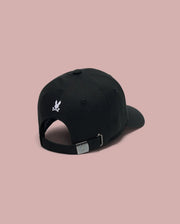 The Psycho Bunny KIDS ARCHIVE BASEBALL CAP (KO1500004) is a black cotton twill cap shown from the back, featuring a small embroidered bunny logo above the adjustable strap with an engraved silver buckle. Background is solid light pink.