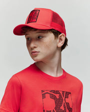 A young boy with light skin and freckles wears the Psycho Bunny KIDS GLENN TRUCKER CAP (KO1500012) and a matching red T-shirt with a black graphic, looking to his left against a plain, light background.