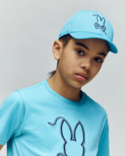 A young person wearing the Psycho Bunny KIDS CYRUS BASEBALL CAP in light blue, featuring a bunny and rope graphic, stands against a plain background, looking slightly to the side.