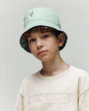 A young boy wears the Psycho Bunny KIDS CHUCK REVERSIBLE BUCKET HAT - KO1500015 in light green, featuring SPF protection, and a cream "Psycho Bunny" sweatshirt, posing against a plain light gray background.