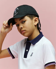 A young person with braided hair wears the Psycho Bunny KIDS ARCHIVE BASEBALL CAP - KO1500004 in black cotton twill and a white polo with navy trim, standing against a pink background while slightly lifting the cap's brim.