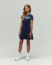 A young girl stands against a plain light background, wearing the Psycho Bunny RETAIL BALL CREW GIRLS DRESS (FR) - KO2000004, paired with white socks featuring a black logo and white athletic shoes with green and blue details.