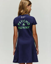 A girl with light brown hair wears the Psycho Bunny RETAIL BALL CREW GIRLS DRESS (KO2000005), a short-sleeved navy blue dress. The back displays "PSYCHO BUNNY" in bright green with a small bunny logo between the words.