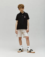 A boy with curly hair poses against a plain background, wearing a black Psycho Bunny KIDS MISCHIEF BUNNY PIQUE POLO (KT0100018) crafted from soft Pima cotton with mother-of-pearl buttons, light shorts, white socks with a black design, and black-and-white sneakers.