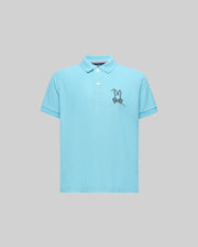 The KIDS CYRUS PIQUE POLO (KT0100031) by Psycho Bunny is a light blue short-sleeve Pima cotton polo with a two-button placket and an embroidered cartoon bunny on a skateboard, shown on a plain gray background.