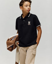 A boy wears the Psycho Bunny KIDS WADE PIQUE POLO (KT0100032) in black, paired with beige cargo shorts and a brown jacket over his arm, standing against a plain light background.