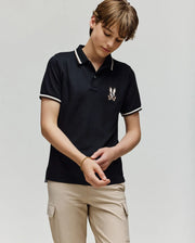 A young person wears the Psycho Bunny KIDS WADE PIQUE POLO - KT0100032 in black with white tipping and beige cargo pants, looking down while adjusting a bracelet against a plain light background.