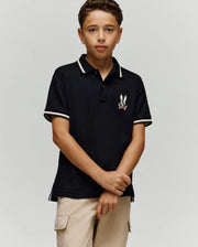 A boy with short curly hair wears the Psycho Bunny KIDS WADE PIQUE POLO - KT0100032 in black with a small embroidered logo, paired with beige cargo pants, standing against a plain light gray background.
