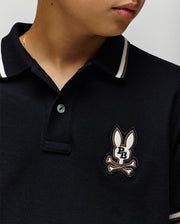 Close-up of a child wearing the Psycho Bunny KIDS WADE PIQUE POLO (KT0100032) in black, featuring white colorblock tipping and an embroidered bunny logo with "PB" letters and crossed bones on the chest. The face is partially visible.