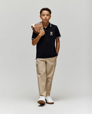 A boy stands against a light gray background, wearing Psycho Bunny’s KIDS WADE PIQUE POLO (KT0100032) in black with an embroidered logo, beige cargo pants, and white sneakers. He holds a brown baseball glove over his shoulder and looks at the camera.