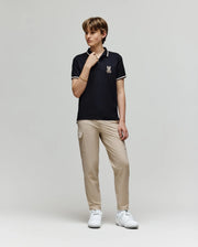 A young person stands against a plain background wearing the Psycho Bunny KIDS WADE PIQUE POLO - KT0100032 in black, featuring an embroidered logo, paired with beige cargo pants and white sneakers, looking to the side with one hand on their collar.