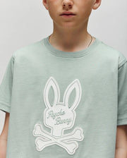 A child wears the Psycho Bunny KIDS DEREK CENTER GRAPHIC TEE (KT0200006), a light green regular-fit shirt featuring a large white bunny head with crossbones and "Psycho Bunny" on the front. Part of the face is out of frame; the background is plain.