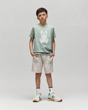 A boy stands against a plain background, wearing the Psycho Bunny KIDS DEREK CENTER GRAPHIC TEE (KT0200006) in light green Pima cotton with an embroidered bunny graphic, beige shorts, white socks with a green logo, and white sneakers featuring green details.