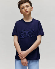 A boy with short brown hair wears the Psycho Bunny KIDS ELIAS GRAPHIC TEE (KT0200013) in navy blue, featuring a retro Bunny graphic, and light blue shorts, standing against a plain light gray background with arms loosely crossed.